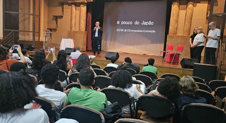 #paratodosverem: a imagem mostra um auditório com vários(as) estudantes sentados(as) em cadeiras estofadas, assistindo a uma palestra.