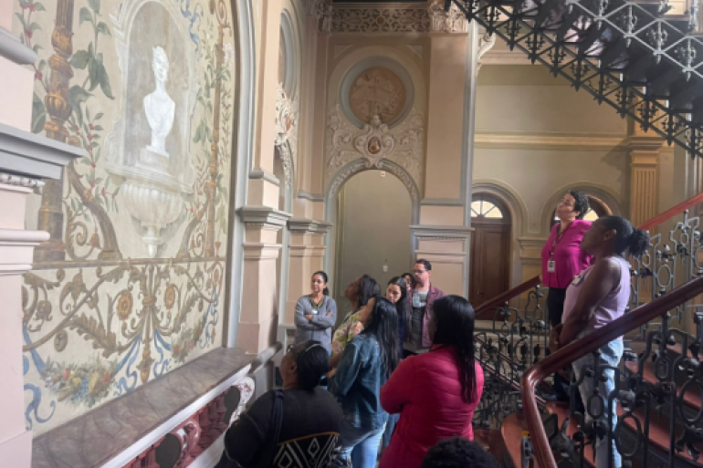 Visita representa uma oportunidade para aprender fora do hospital. #paratodosverem: Um grupo de visitantes no Palácio da Liberdade observa uma grande pintura mural à esquerda, que retrata um busto branco em um nicho ornamentado. As pessoas, em sua maioria mulheres, estão distribuídas entre o piso inferior e os degraus de uma imponente escadaria de ferro trabalhado à direita. O ambiente possui arquitetura clássica com arcos, colunas e teto decorado.