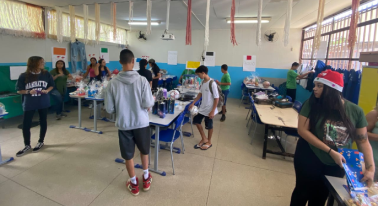 #paratodosverem: a foto mostra uma sala com paredes pintadas em azul claro e branco, decorada com tiras de papel penduradas no teto. Há várias mesas organizadas em fileiras, sobre as quais estão expostos diversos produtos, como bolas de futebol, fones de ouvido, doces, calçados, itens de maquiagem e kits embrulhados em celofane. Vários(as) estudantes circulam pela sala, observando os itens. 