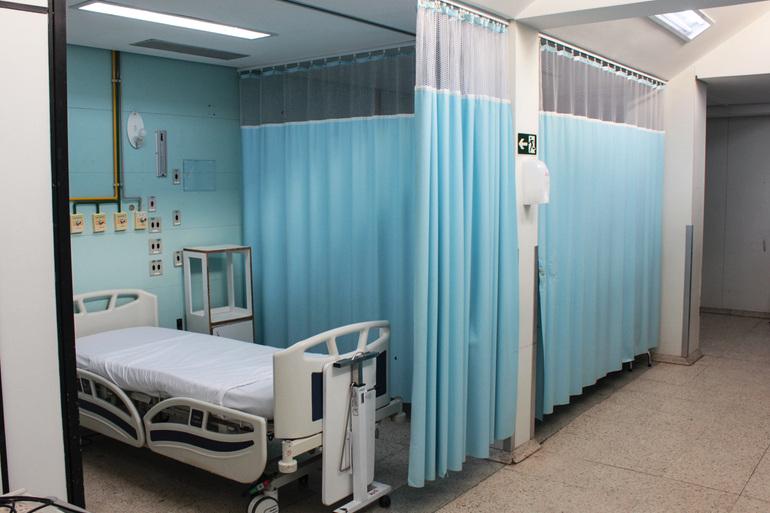 Leito de hospital com uma cama hospitalar branca vazia e aparelhos de apoio, um leito com cortinas azuis abertas e outro com cortinas fechadas. 