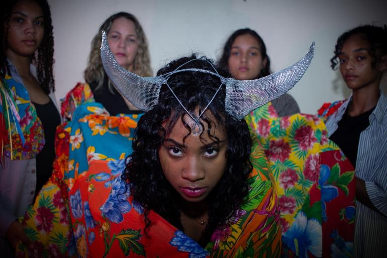 Espetáculo promove encontro entre memória, tradição e contemporaneidade no Teatro Marília Fotografia colorida em plano médio de cinco mulheres. No centro, em destaque, uma jovem mulher negra com cabelos cacheados escuros olha fixamente para a frente, com a cabeça levemente inclinada para baixo e um olhar intenso.