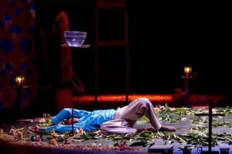 Cena do espetáculo teatral "Capelas Gerais". Um ator está deitado de costas no chão do palco, cercado por folhas verdes e flores espalhadas. Ele veste blusa azul de mangas compridas e calça clara. Acima dele, há uma tigela de vidro com água equilibrada sobre um suporte alto. Ao fundo, o cenário escuro traz velas acesas e uma cruz no canto inferior direito.