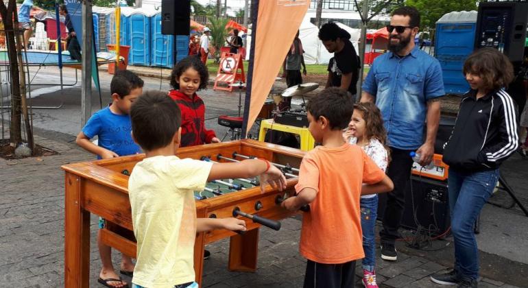 Quatro crianças brincam de pebolim, acompanhadas por uma criança e um adulto, durante o dia.