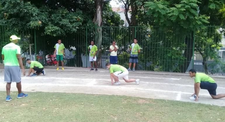 Técnico acompanha três atletas corredores em suas marcas, na pista de corrida, enquanto outras quatro pessoas observam. 