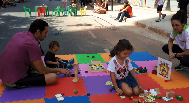 Duas crianças brincam com jogos diversos, no chão, acompanhadas por adulto.