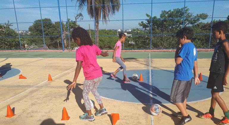 Quatro crianças têm aulas de futsal em quadra, durante o dia.