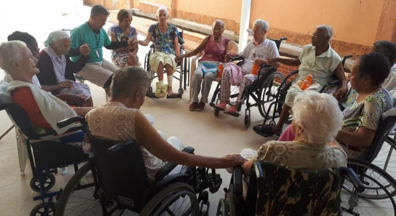 Onze idosos cadeirantes em círculo e de mãos dadas com um instrutor. 