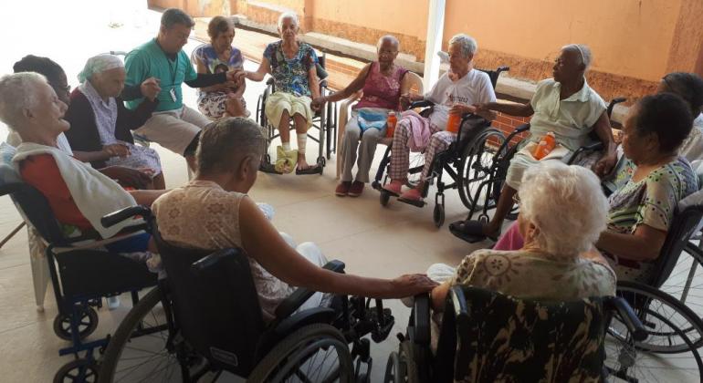 Programa da Prefeitura de Belo Horizonte alegra a rotina dos residentes de Instituições de Longa Permanência para Idosos. Foto: Vilma Tomaz/PBH Onze idosos cadeirantes em círculo e de mãos dadas com um instrutor.
