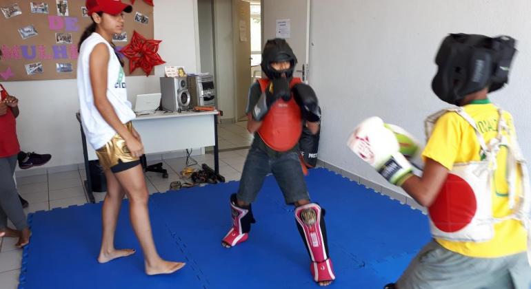 Dois alunos devidamente equipados praticam Muay Thai sob supervisão da professora, em um tatami.
