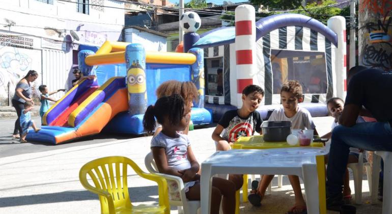 Quatro crianças sentadas próximas a uma mesa de plástico; ao fundo, três crianças brincam em escorregador de plástico, acompanhadas por adulta. 
