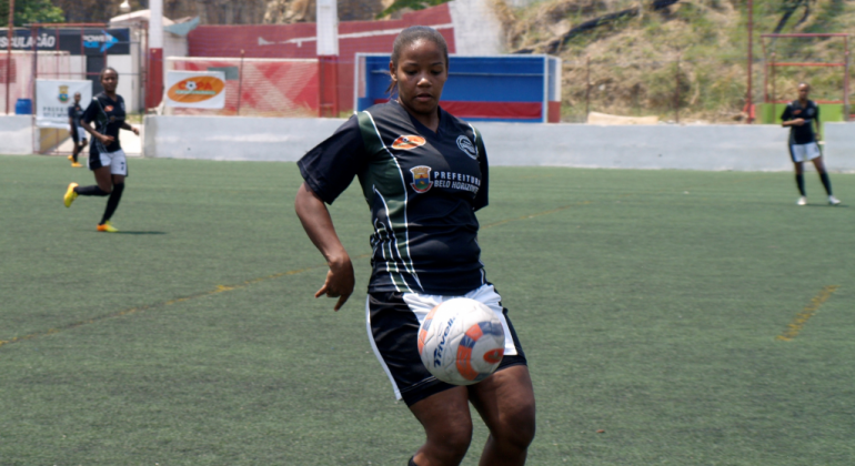 Jogadora toca a bola em campo de futebol, durante o dia. 