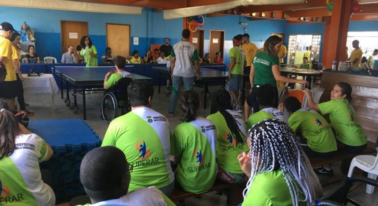 Mais de sete crianças observam campeonato paralímpico de pingue-pongue em uma sala. 