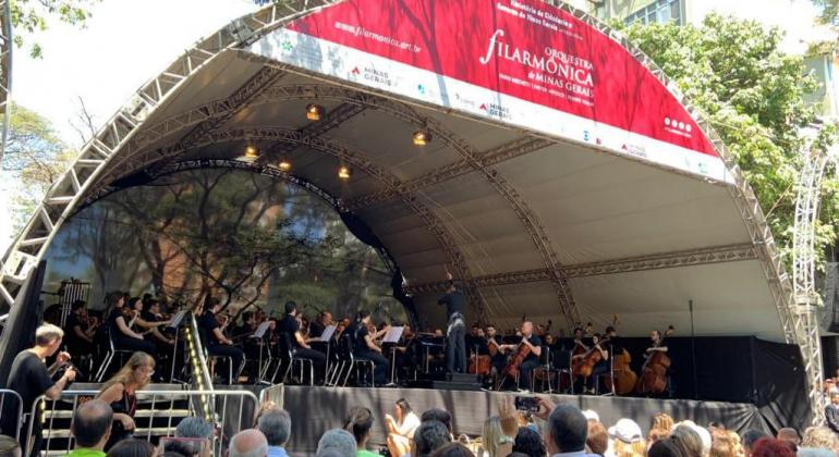 Apresentação da Orquestra Filarmônica na Savassi. AO fundo aparece toda a orquestra e à frente parte do público