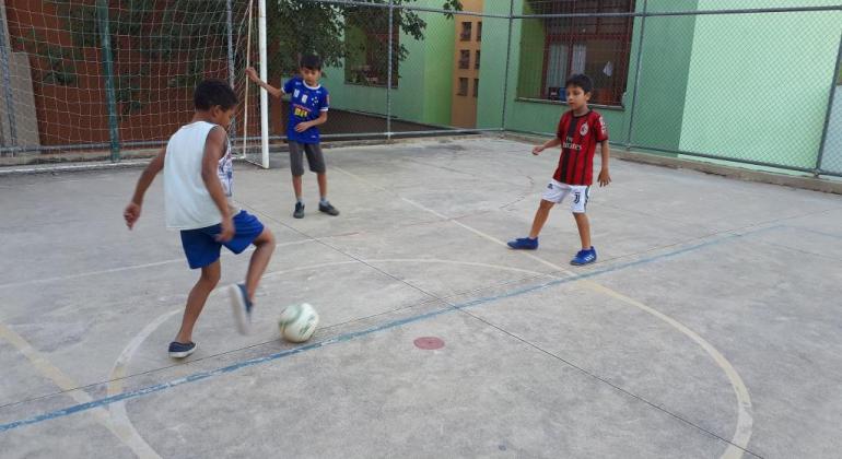 Três garotos jogam futebol em quadra, durante o dia. 