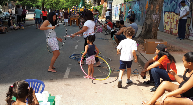 Show da Banda Travessia é a atração do BH é da Gente - unidade Guarapari; jogos infantis são o destaque da unidade Silva Lobo. Foto: Divulgação PBH Três crianças brincam de bambolê no BH é da Gente - av. Silva Lobo; ao redor, pessoas acompanham a brincadeira e passeiam.