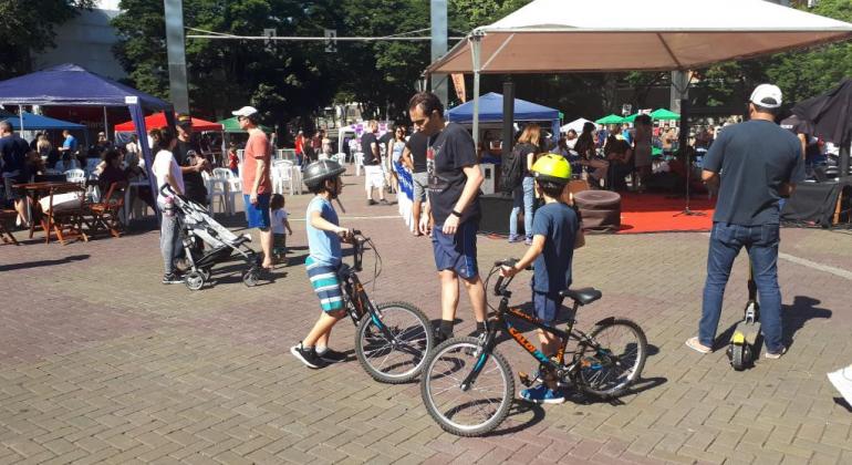 Duas crianças estão de pé, segurando suas bicicletas, acompanhadas por adulto, também de pé. Ao fundo, pessoas passeiam, durante o dia. 