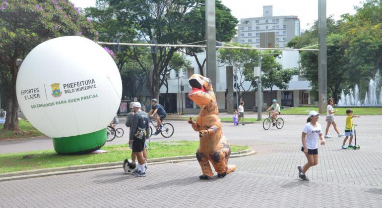 Homem fantasiado de dragão caminha em rua fechada, ao lado de bola com a logo da Prefeitura; pessoas caminham ao redor, durante o dia. 