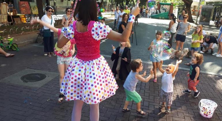 Mulher com vestido de bolinha faz bolhas de sabão, enquanto as crianças sorriem e tentam agarrá-las. 