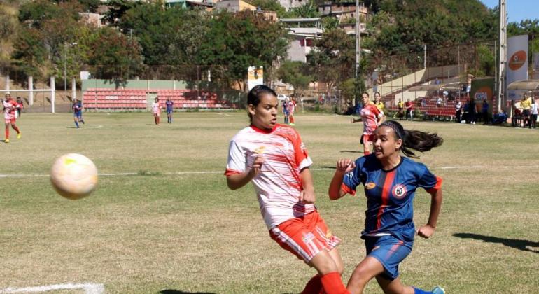 Duas garotas de times diferentes jogam futebol em campo; à direita, a bola está no ar. 