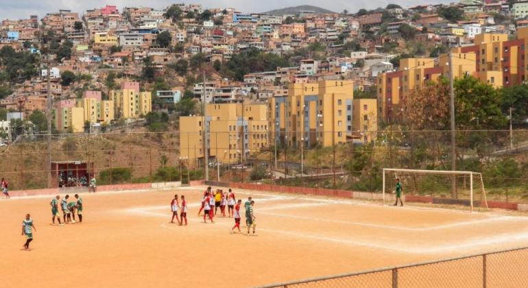 Times jogam em quadra de futebol de sem grama com cidade ao fundo. 