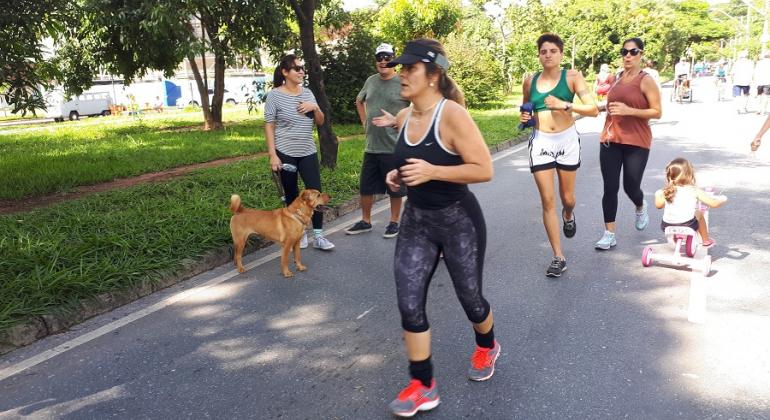 Três mulheres fazem corrida em avenida durante o dia; ao fundo, criança brinca e mulher passeia com cachorro. 