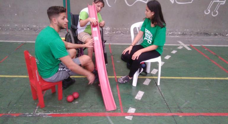 Aluno com deficiência joga bocha, auxiliado por dois adultos e uma prancha, por onde a bola desliza. 