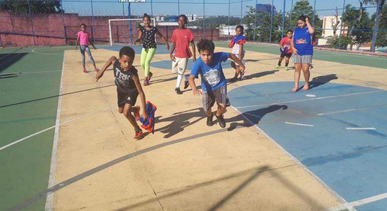 Oitro crianças jogam futebol em quadra poliesportiva a céu aberto, durante o dia. 