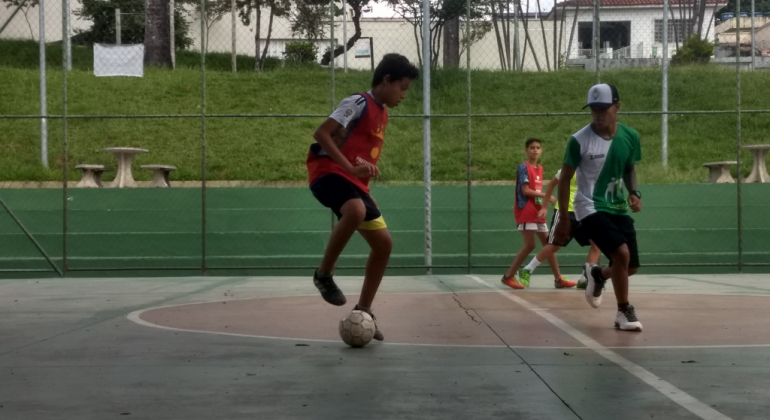 Três garotos e uma bola em quadra poliesportiva, durante o dia. 