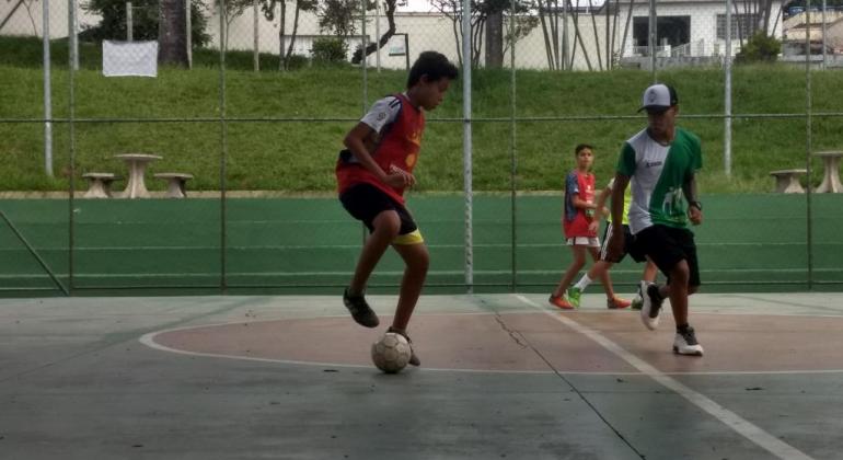 Três crianças jogam futebol em quadra, durante o dia. 