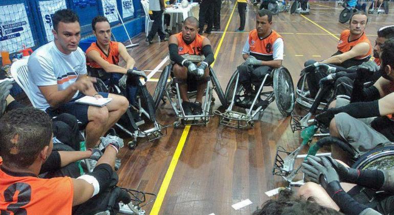 Time de basquete composto por cadeirantes se reúne em círculo e acompanha a orientação de técnico. 