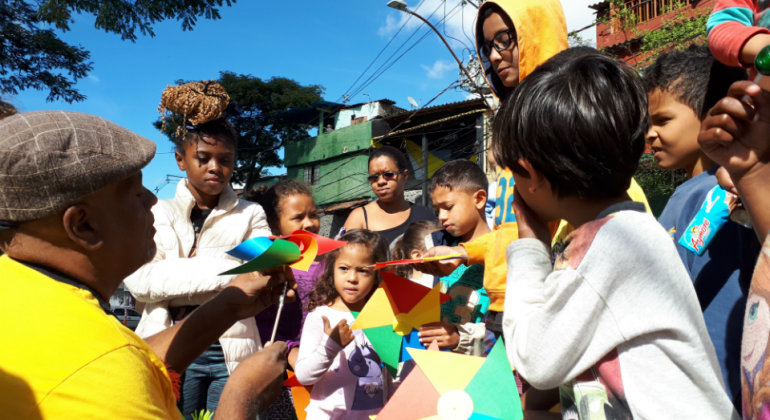 Sete crianças manuseiam três cataventos coloridos, acompanhadas de dois adultos, durante o dia. 