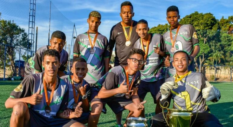 Sábado, dia 6/7, é dia da semifinal do módulo Juvenil da Copa Centenário 2019, com jogos nas regiões Nordeste e Pampulha. Foto: Glass Fotografia Foto de nove jogadores do time Itamarense, com medalhas e taça de campeões do módulo juvenil masculin da Copa Centenário 2018