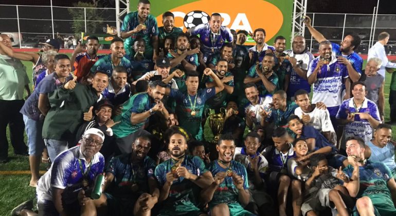 Mais de trinta pessoas, entre membros de um time de futebol e torcedores, comemoram a vitória de uma categoria da Copa Centenário. 