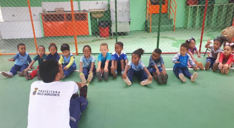 Onze crianças sentadas no chão, ouvindo orientações de rapaz com a camiseta da Prefeitura Municipal de Belo Horizonte. 