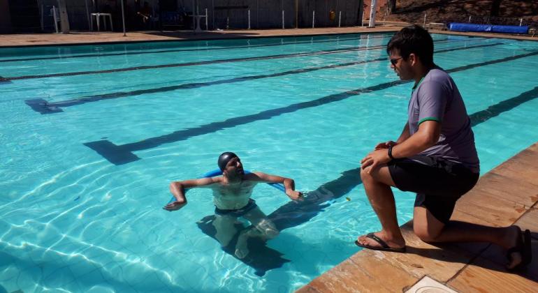 Universitário em piscina com raia olímpica, acompanhado por rapaz fora da piscina, durante o dia. 