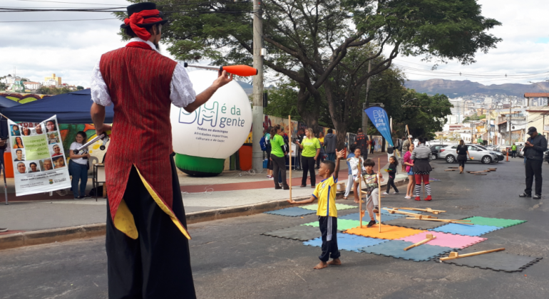 Homem com perna de pau se apresenta para crianças, ao fundo, bola com os dizeres BH é da Gente. 