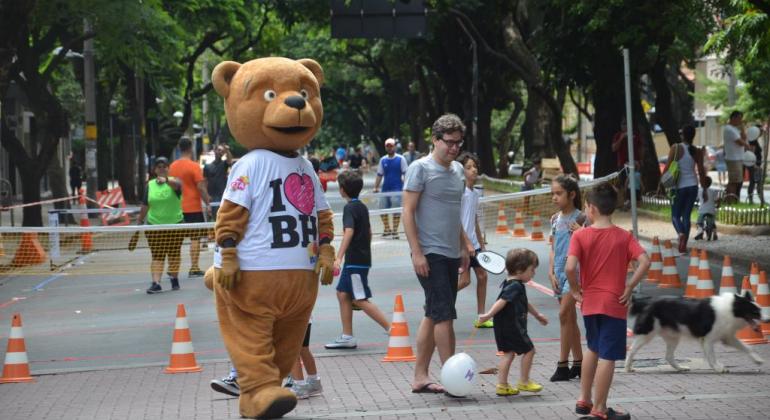 Homem fantasiado de urso com camiseta com os dizeres: "I love BH" passeia em rua com crianças brincando acompanhadas por adulto, durante o dia. 