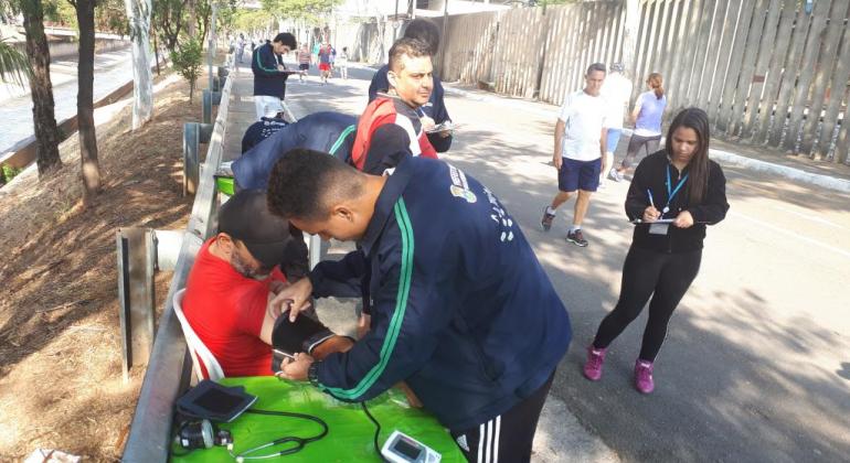 Técnico mede a pressão de cidadão enquanto técnica preenche formulário, ao fundo, pessoas fazem caminhada, durante o dia. 