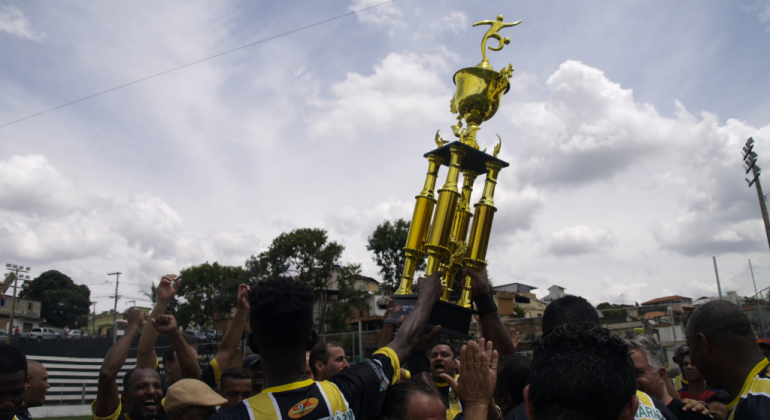 Jogadores de futebol erguem taça comemorativa, durante o dia. 