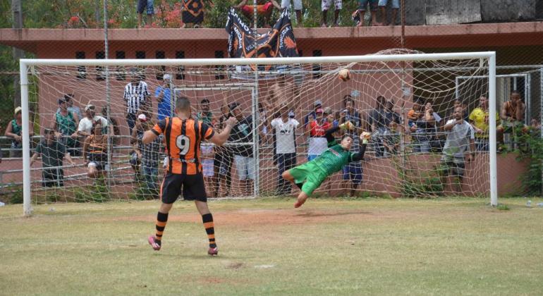 Jogador, de uniforme laranja e preto, dá chute a gol, enquanto goleiro de verde pula no ar para pegar a bola. 
