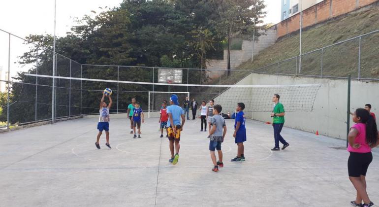 Time misto joga vôlei em quadra poliesportiva descoberta, durante o dia. 