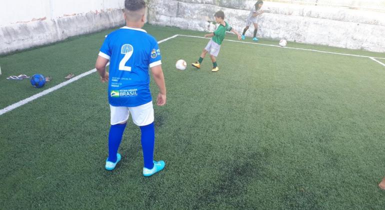 Homem de uniforme de futebol joga bolo com criança, também de bermuda e tênis, em quadra gramada, durante o dia. 