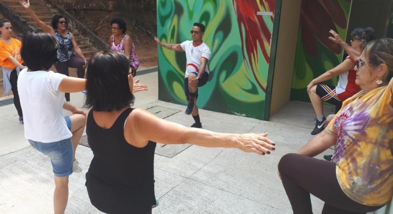 Caminhada orientada, dança, jogos de mesa, karaokê e jogos teatrais fazem parte das ações de férias do Centro de Referência da Pessoa Idosa. Foto: Vilma Tomaz/PBH Instrutor, de pé, faz pose de alongamento com mão esquerda sobre a perna esquerda, dobrada, e é seguido por cerca de seis pessoas, em local fechado.