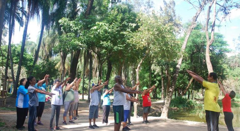 Cerca de dez pessoas fazem ginástica em contato com a natureza, orientadas por dois técnicos da Prefeitura. 