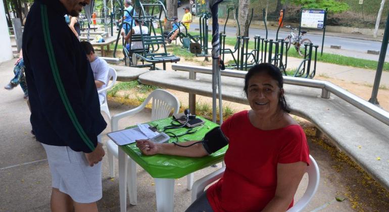 Denise conta com o auxílio do programa Caminhar, desenvolvido pela Secretaria Municipal de Esportes e Lazer. Foto: Marcelo Machado/PBH Mulher sentada mede pressão com técnica de saúde.