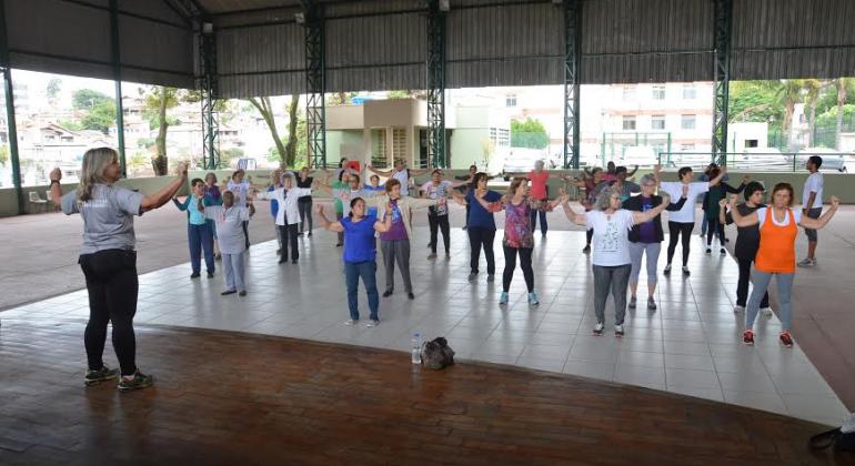 Mulher orienta ginástica de mais de 15 idosos em galpão, durante o dia. 