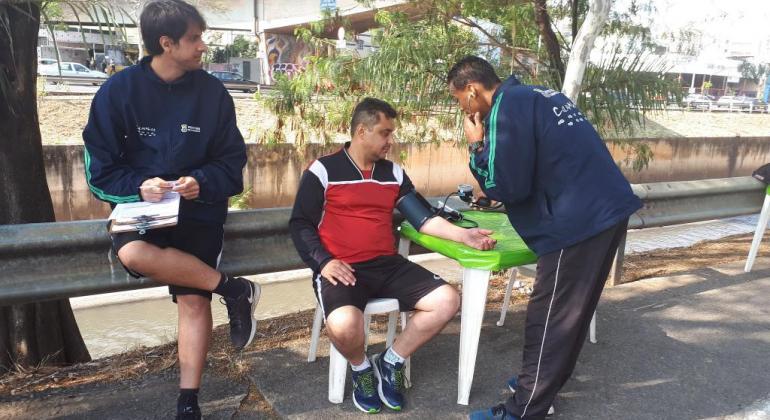 Praticantes de caminhada podem ter acesso a testes de a orientações sobre a forma mais segura de praticar atividades físicas. Foto: Vilma Tomaz/PBH Profissional afere pressão de esportista, sentado, outro faz anotações em uma palheta, durante o dia.