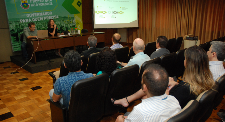 Foto: Divulgação PBH Homem observa laptop e mulher palestra a cerca de oito pessoas sentadas, em uma sala com telão.