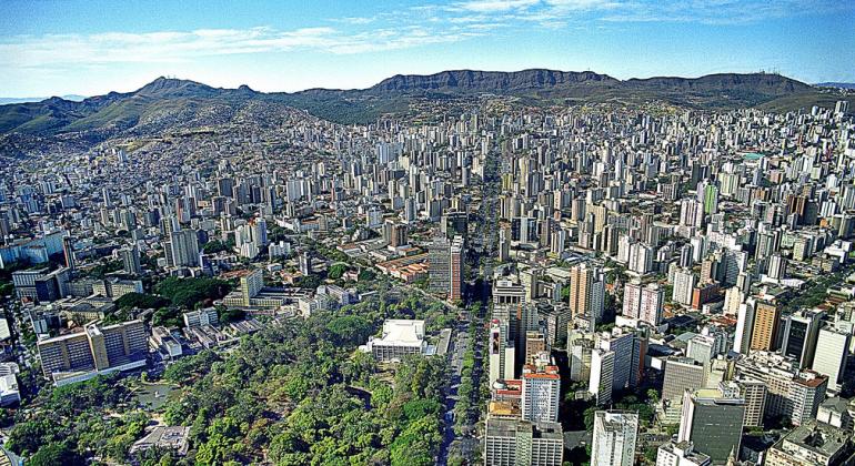 Vista aérea da avenida Afonso Pena, com Parque Municipal à esquerda, abaixo, prédios por todos os lados e a Serra do Curral ao fundo. 