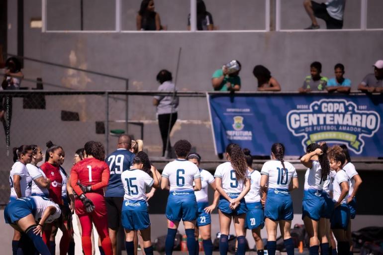 Futebol feminino ganha destaque na rodada da Copa Centenário deste domingo (23) Em um campo de futebol, jogadoras de uma equipe feminina estão reunidas próximas à lateral do gramado, durante uma pausa da partida. A maioria veste camisa branca, shorts azuis e meiões azul-escuros; duas goleiras usam uniformes diferentes, um vermelho e outro azul-marinho.